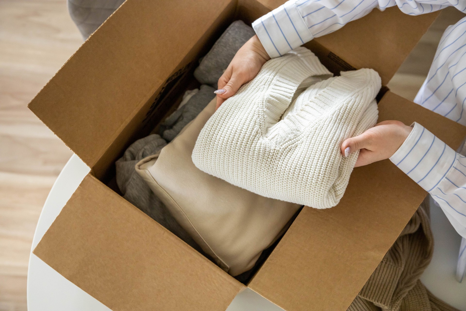 Woman hands putting warm neutral sweater into cardboard box seasonal storage method and donation